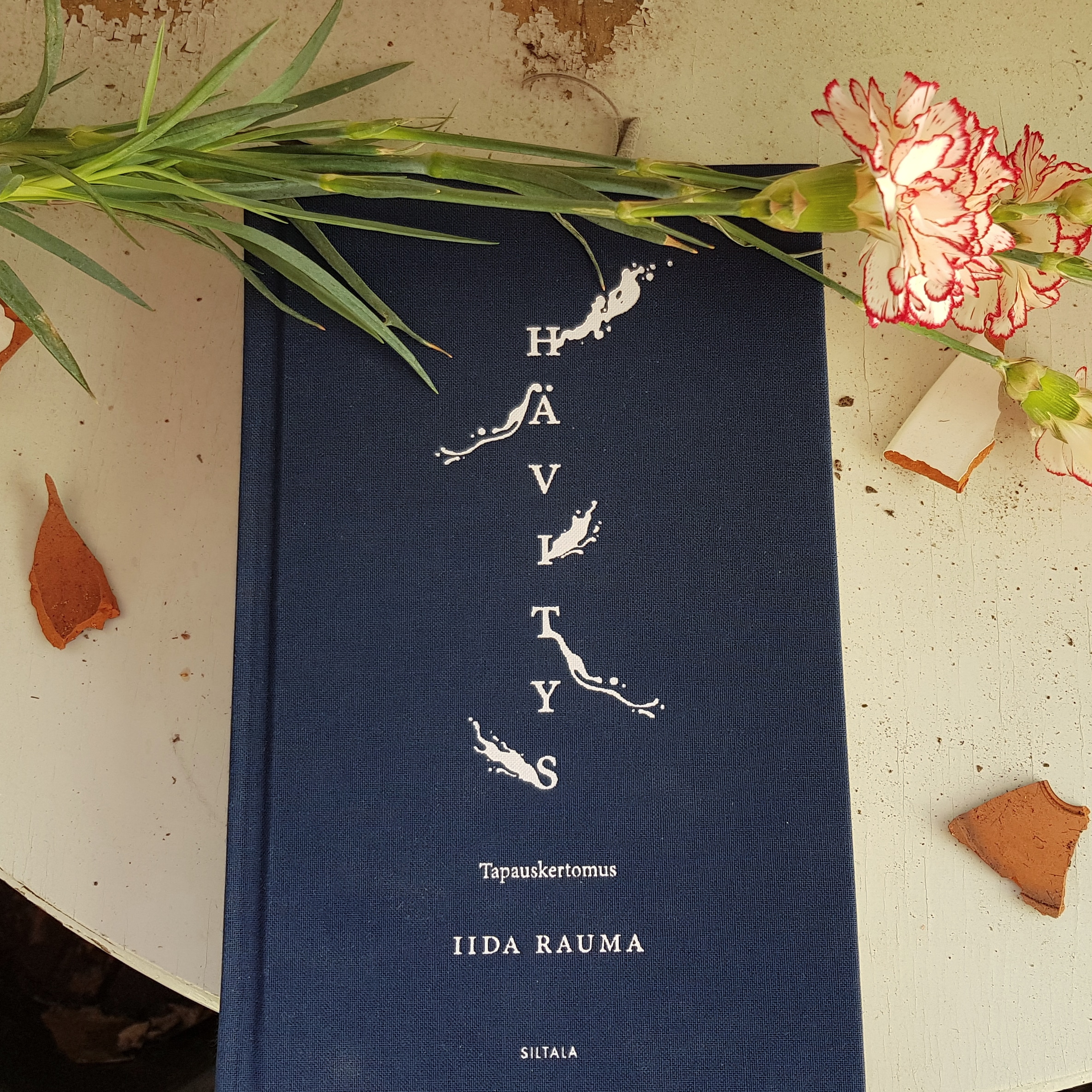 [Kuvassa on kirja vaalean kuluneen pöydän päällä. Sen ympärillä on saven sirpaleita ja punavalkoisia neilikoita. Kirjassa on tummansininen kansi, jossa lukee valkoisella kirjan tiedot. Kirjan nimi Hävitys on kirjoitettu allekkaisin kirjaimin, joista leviää tulenlieskan näköiset tahrat. Sen alla lukee "Tapauskertomus" ja kirjailijan nimi Iida Rauma sekä kustantaja Siltala.]